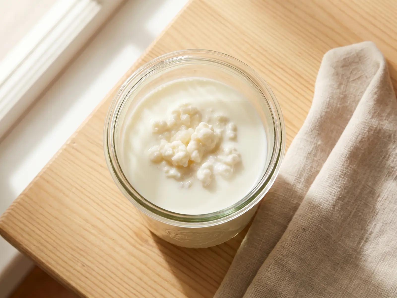 Glas mit fermentierendem Milchkefir auf Holz, daneben ein Leinentuch in ruhiger K&uuml;chenansicht.