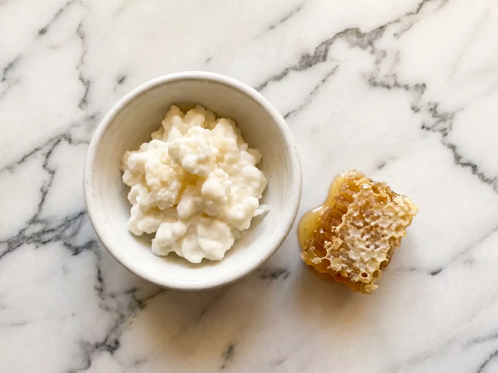 Schale mit cremigem Milchkefir neben einer Honigwabe auf hellem Marmor in warmer Aufsicht