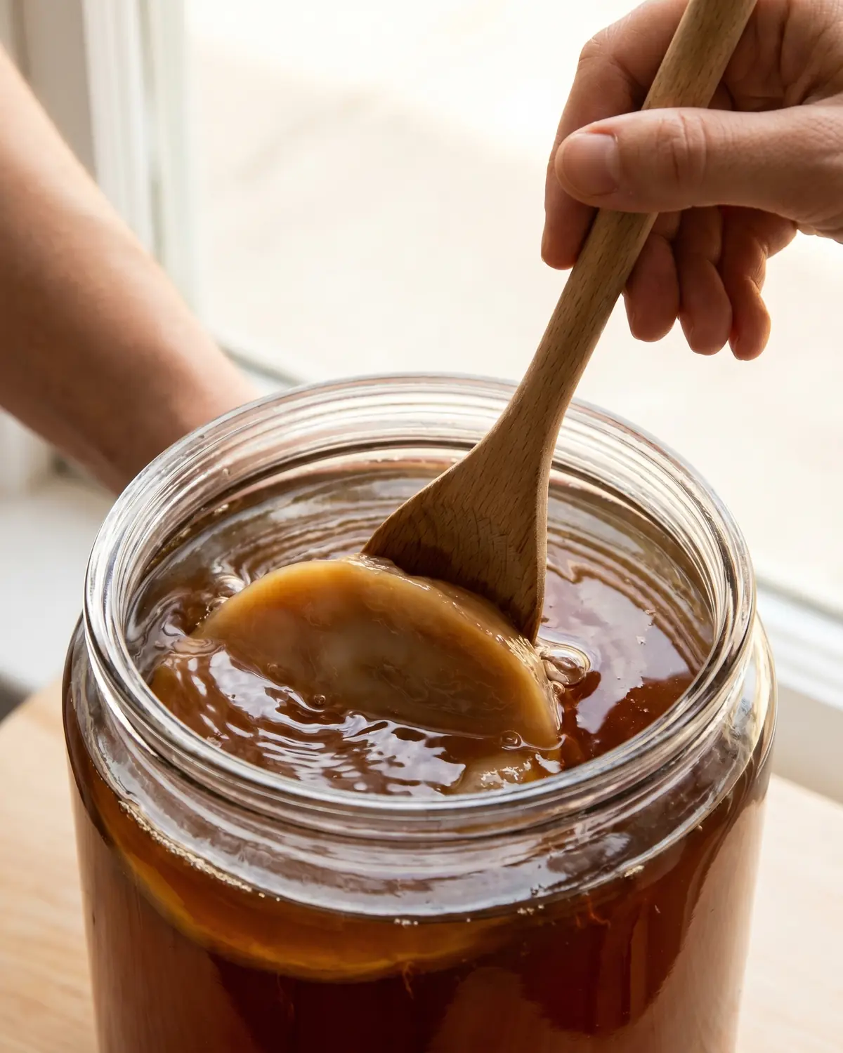 Eine Holzl&ouml;ffelspitze dr&uuml;ckt den SCOBY vorsichtig unter bernsteinfarbenen Tee in einem Glasgef&auml;&szlig;.