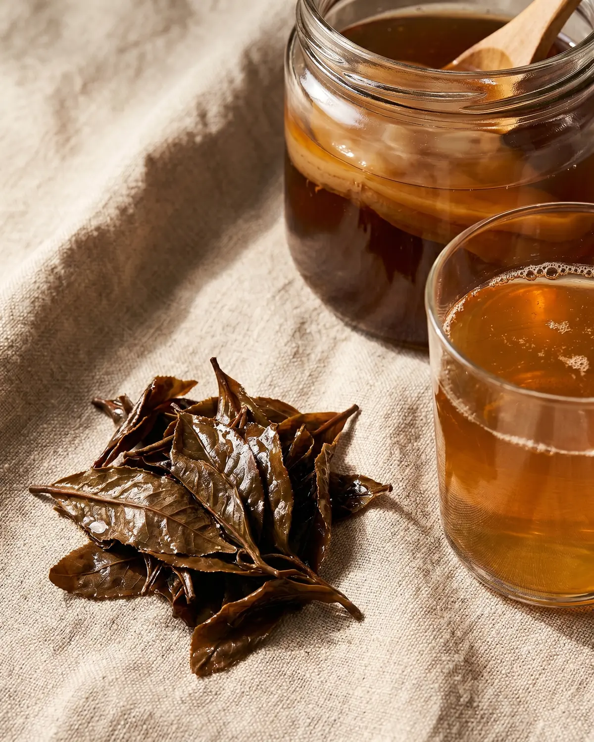 kombucha alkoholgehalt Feuchte Teeblätter liegen neben einem Glas Kombucha auf rohem Leinen und zeigen die Teebasis.
