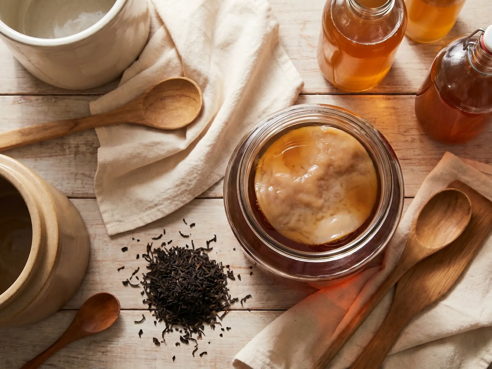 Blick von oben auf Kombucha mit SCOBY, Baumwolltuch, Teebl&auml;ttern und hellem Holz in ruhiger K&uuml;chenstimmung