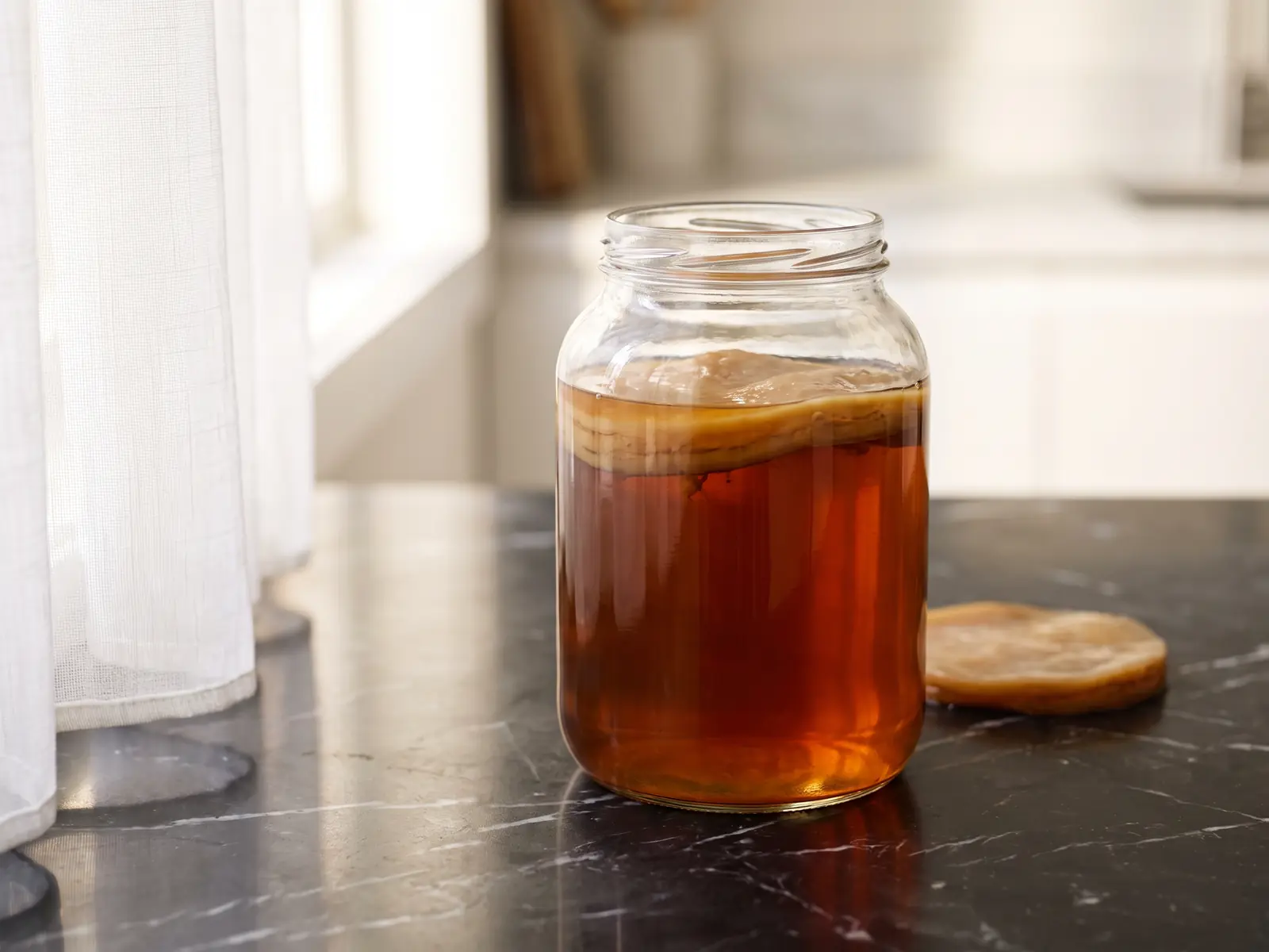 Glas mit SCOBY und tief bernsteinfarbenem Tee auf mattem schwarzem Marmor mit hellem Vorhangspiegelbild.