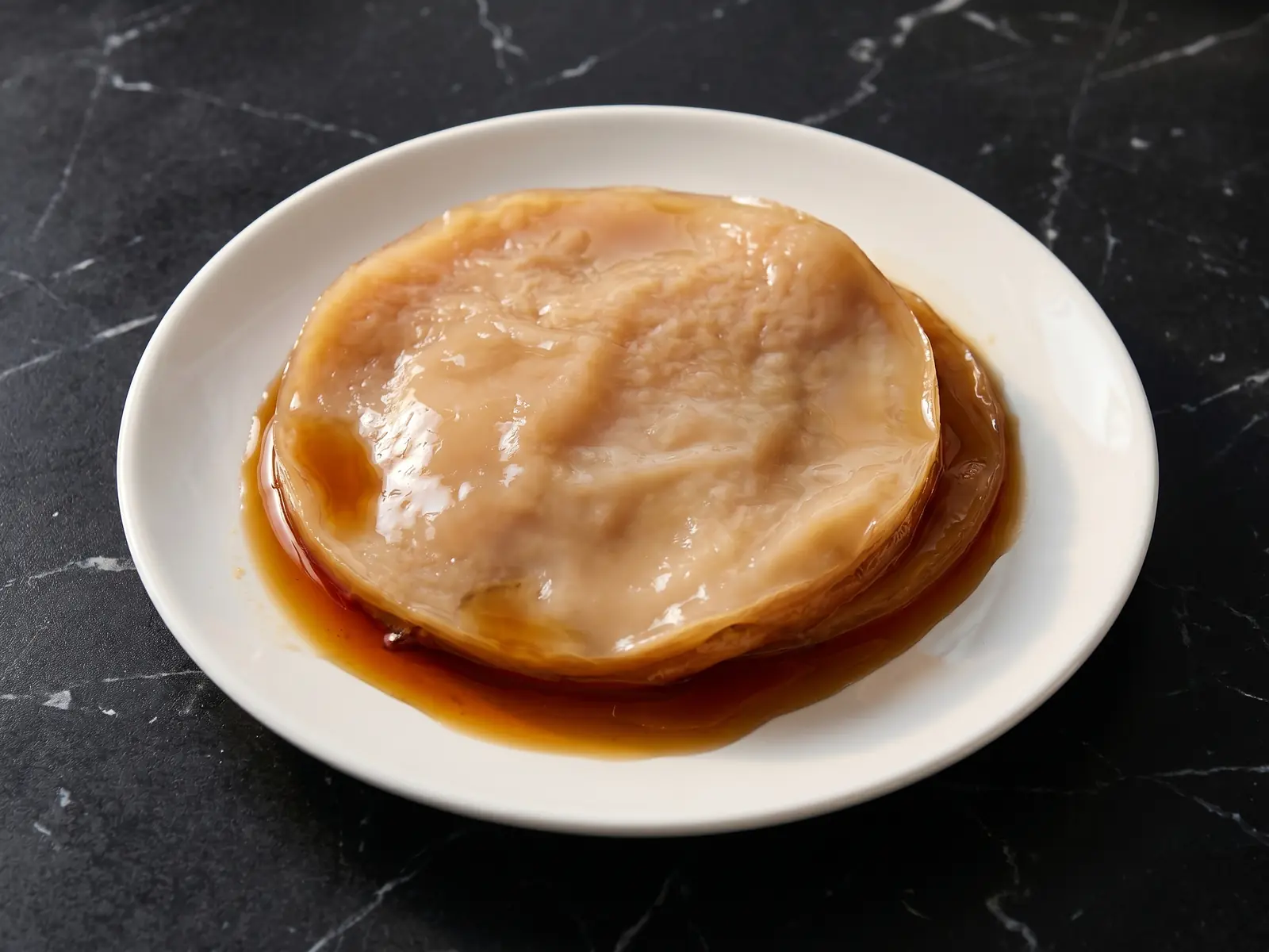 scoby schimmel Beige-brauner Kombucha-SCOBY auf einem weißen Teller, mit feuchter Oberfläche auf dunklem Marmor.