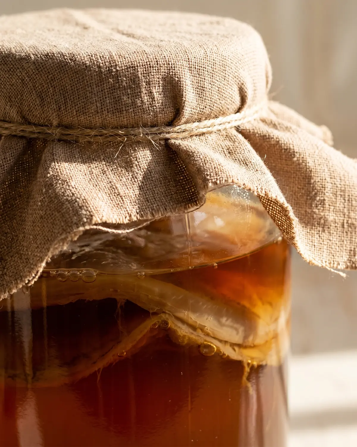 kombucha oder kefir Nahaufnahme eines mit Baumwolltuch bedeckten Kombucha-Glases, dunkler Tee ist darunter sichtbar.