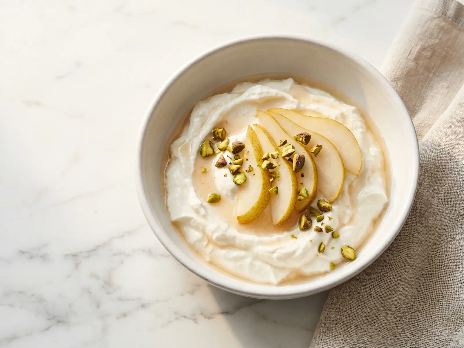 joghurt selber machen Cremiger Joghurt in einer weißen Keramikschale auf Marmor, garniert mit Birne und Pistazien.