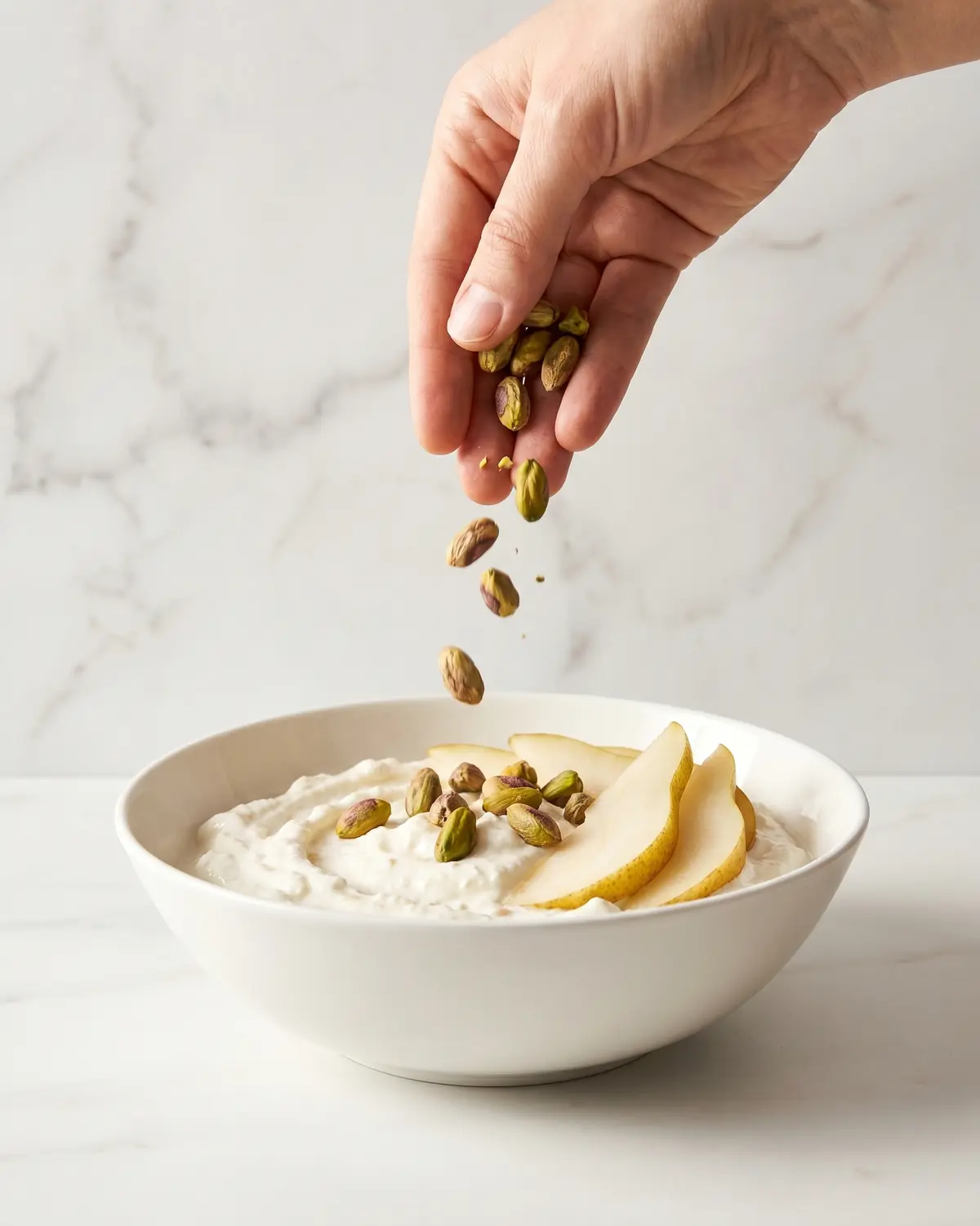 Eine Hand streut Pistazien &uuml;ber Joghurt mit Birne in einer Schale auf hellem Marmor.