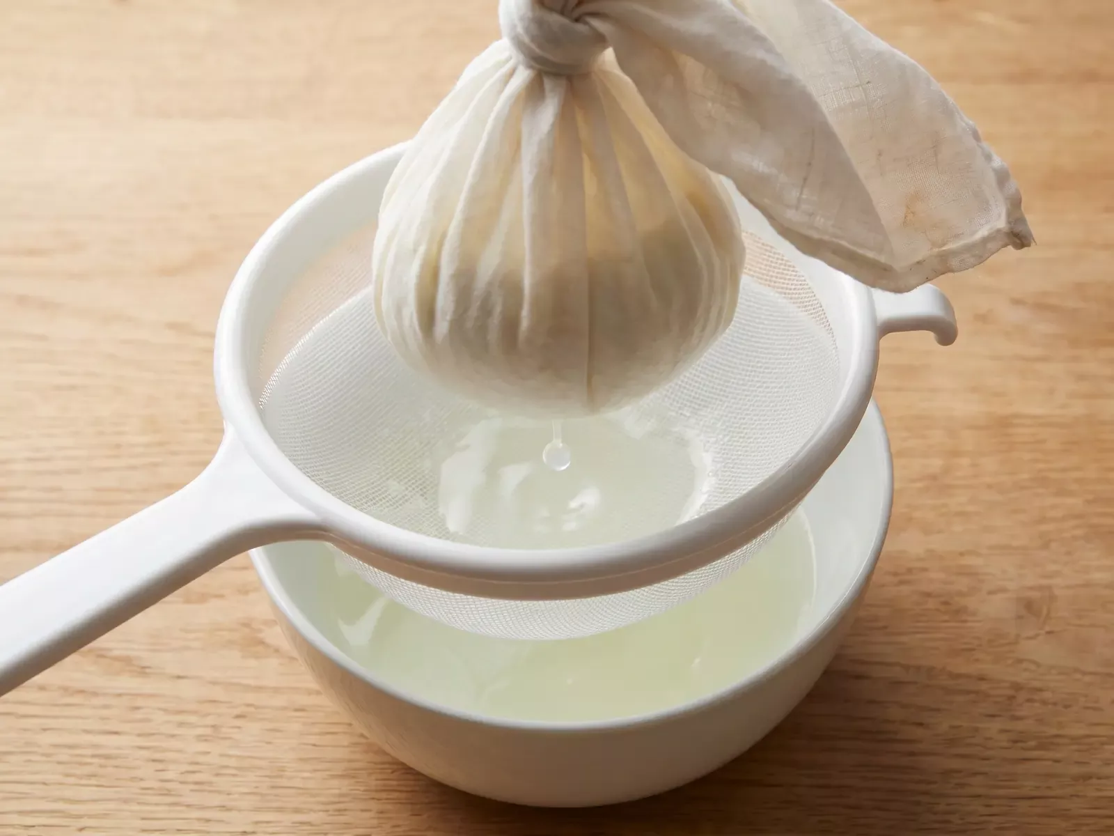 Abgetropfter Joghurt in einem mit Tuch ausgelegten Sieb, w&auml;hrend ein Tropfen Molke herabf&auml;llt.